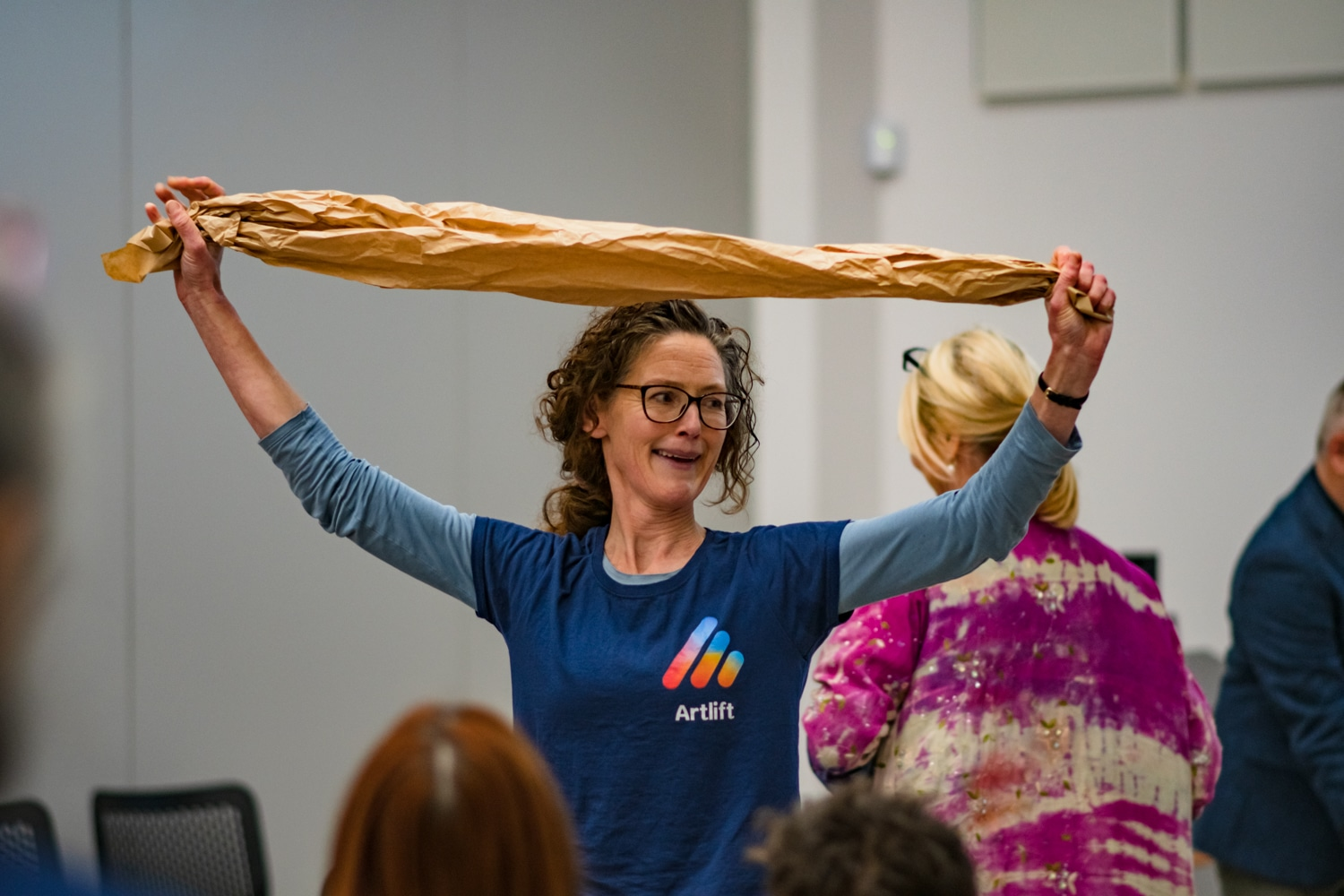 A white woman with brown hair and glasses is holding up some brown paper above her head with both hands and smiling at people in a room during a creative workshop